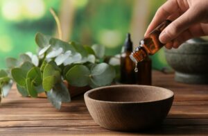 Hand holding an essential oil bottle placing a drop into a wooden bowl next to leaves on a table